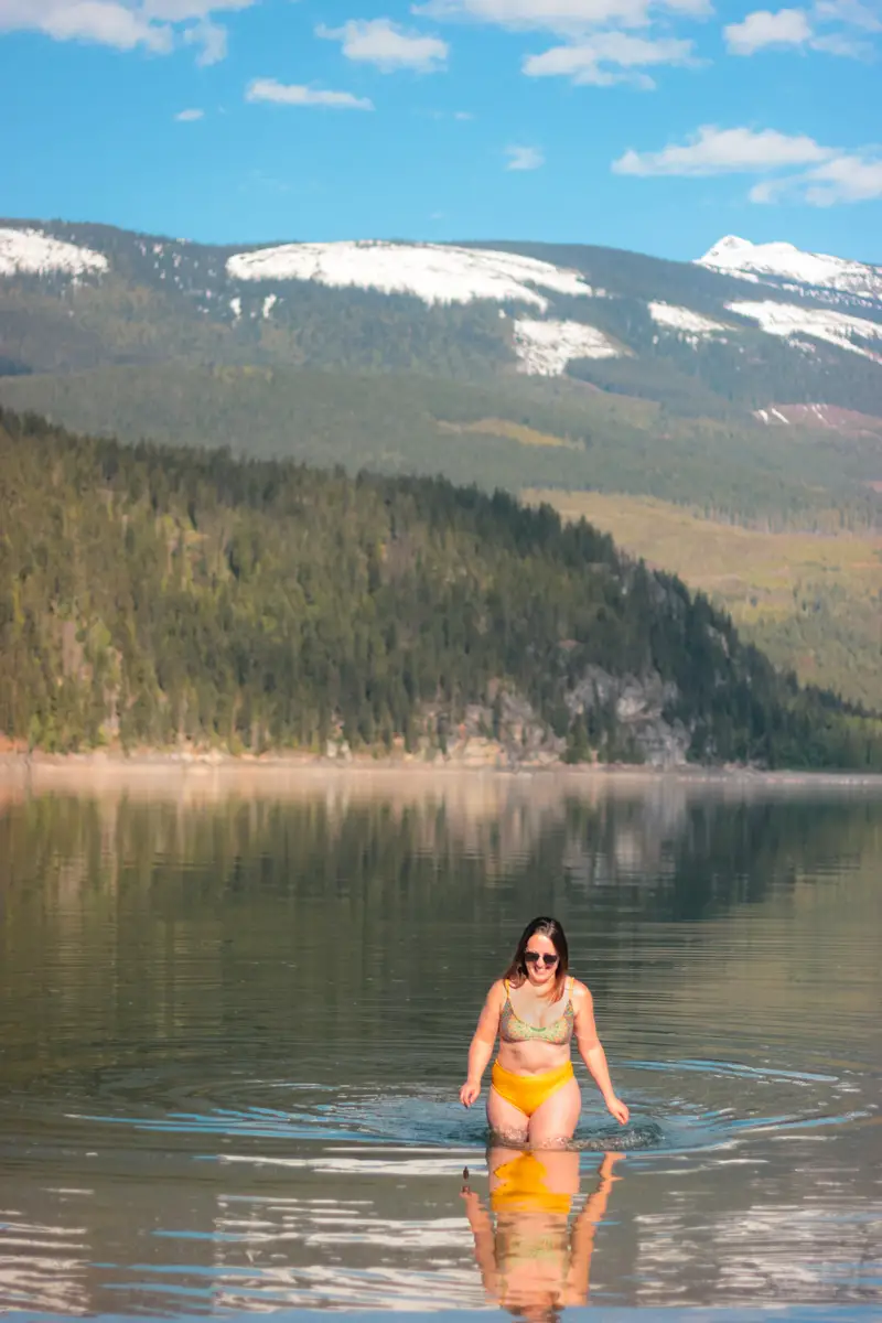 Woman in yellow bathing suit walks out of Arrow Lake with snow capped mountain in the background.