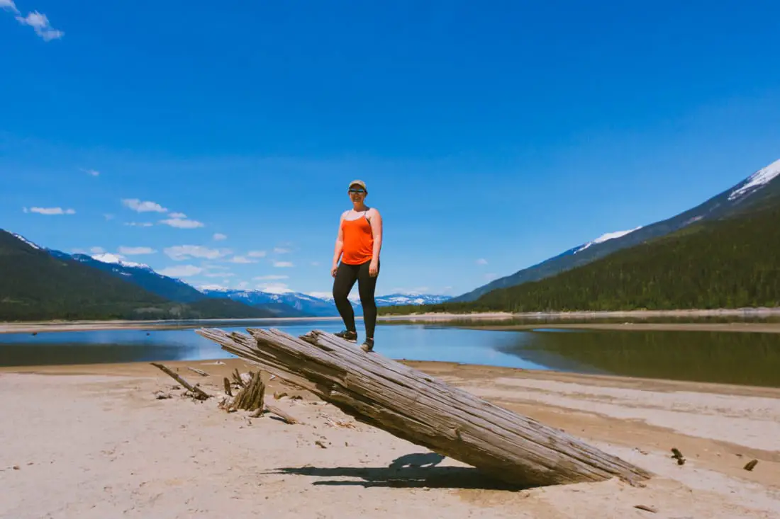 Woman in red tank top stands on top of a large piece of driftwood on the shores of Arrow Lake. Sunny day.