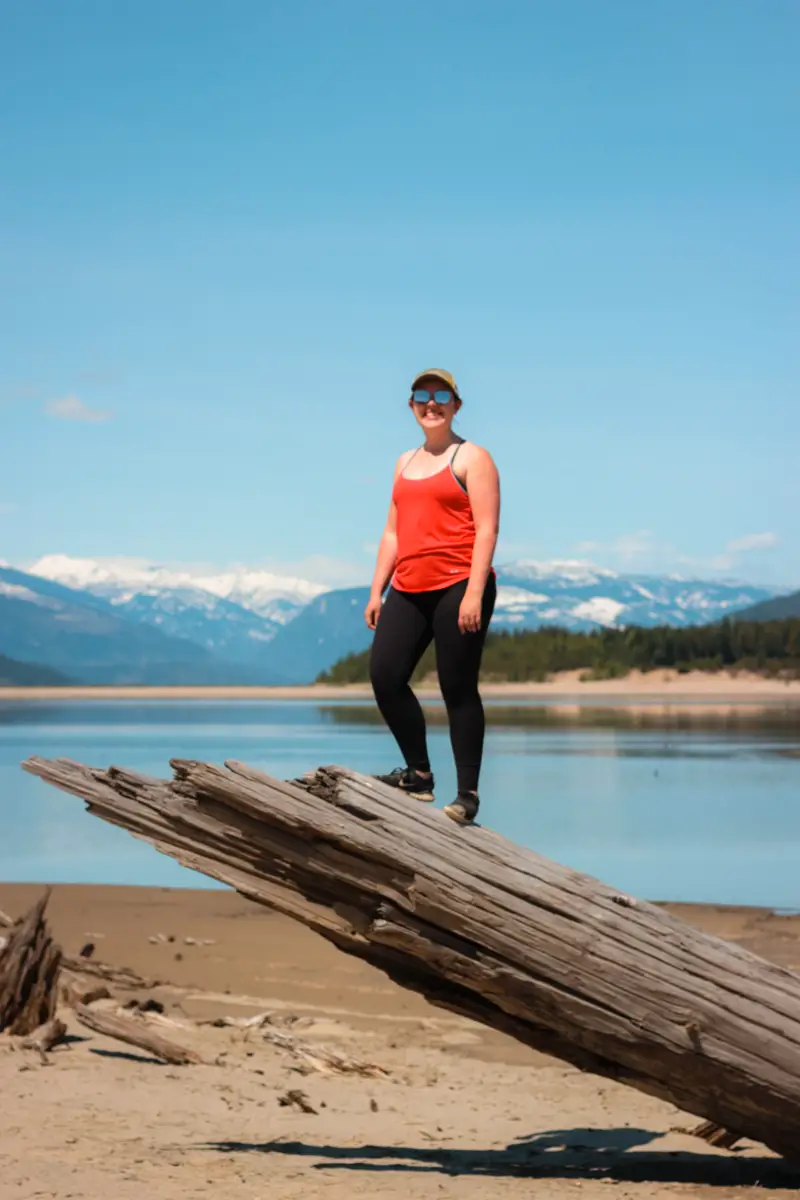 Woman in red tank top and hat stands on a large piece of driftwood on Arrow Lake.