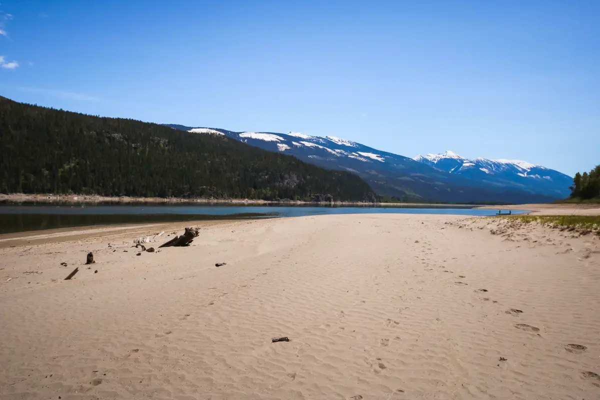 Large, sandy beach on the shores of Arrow Lake with snow capped mountains in the background.