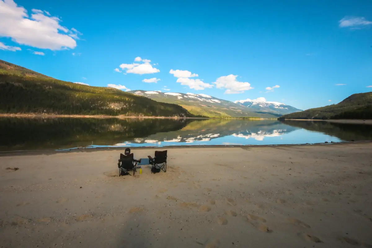 Man sits in one of two camp chairs on a sandy beach on Arrow Lake near Revelstoke BC. Sunny day with blue skies.