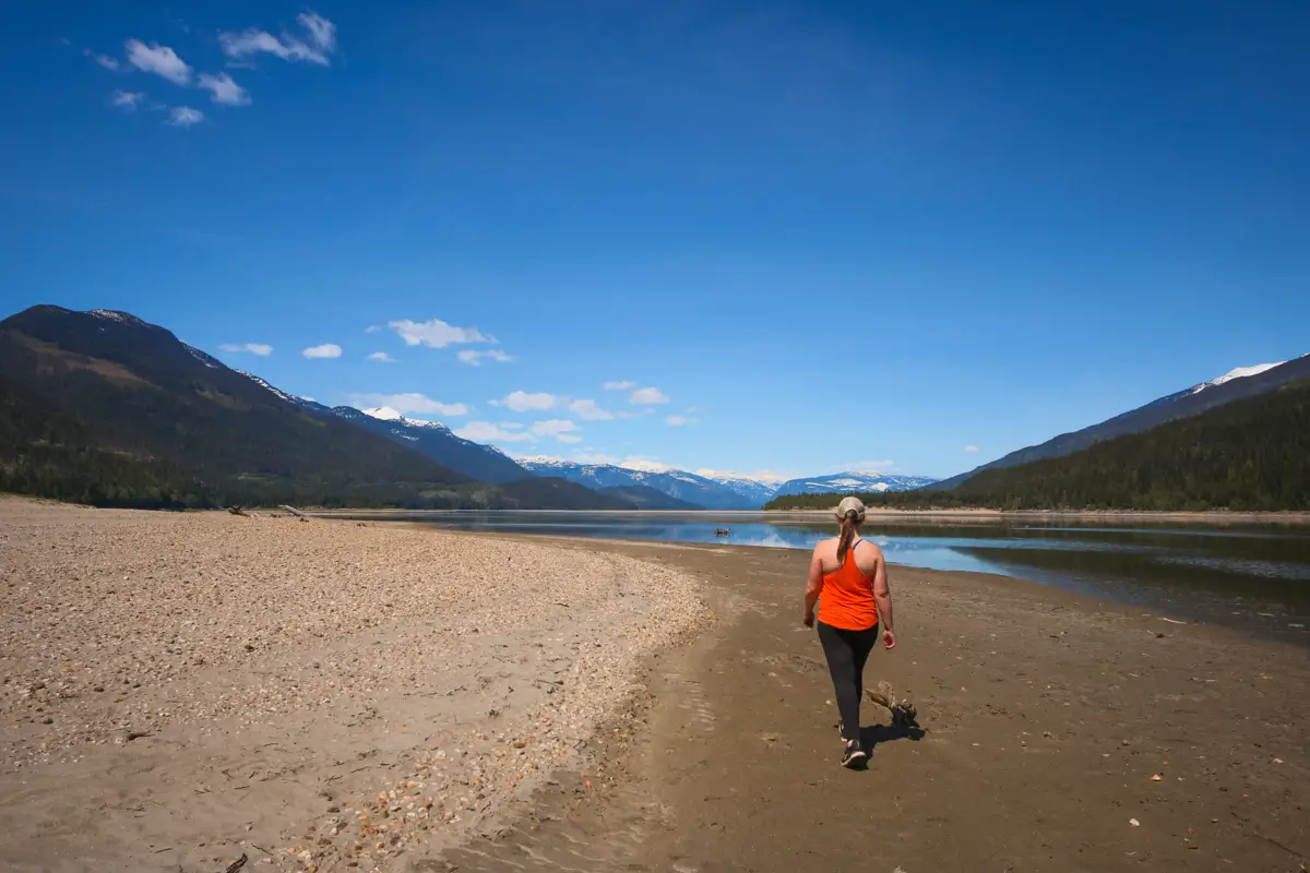 Woman in red walks down a sandy beach on Arrow Lake. Sunny day and blue skies.