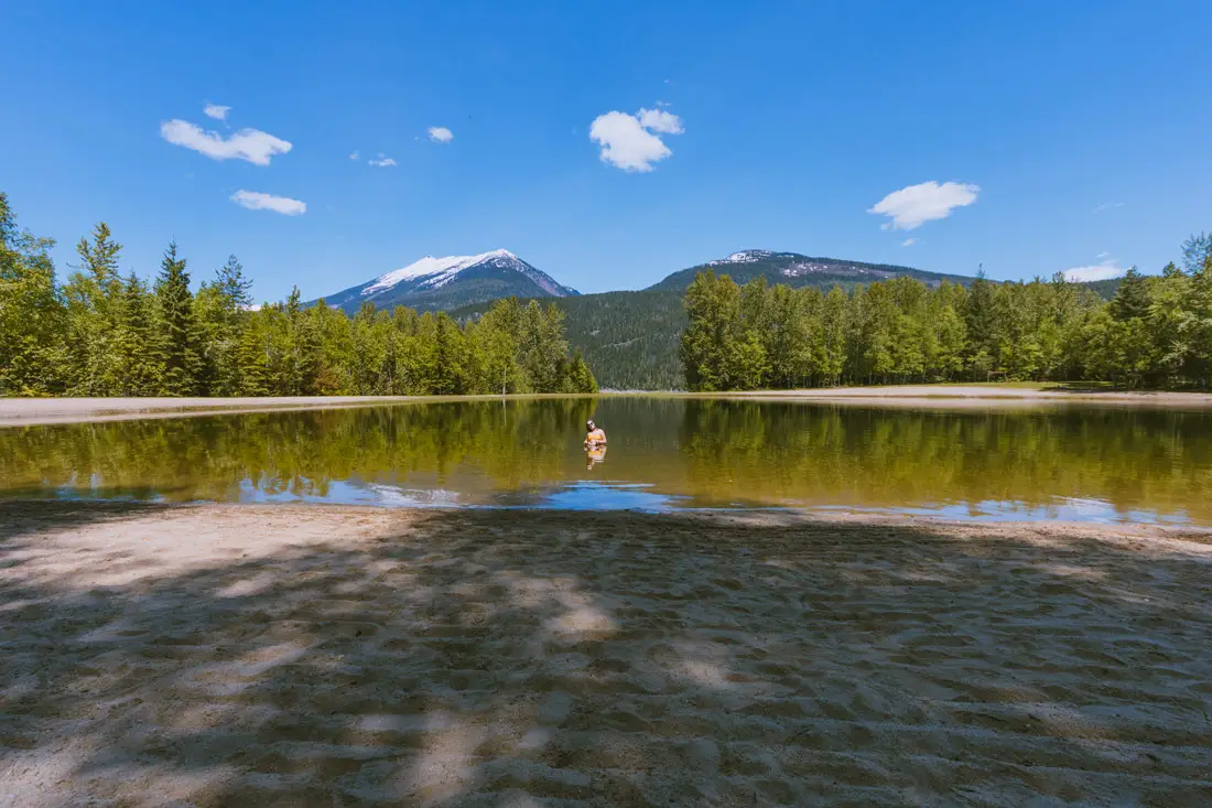 Woman swimming in a sandy, clear lagoon at Blanket Creek Provincial Park. Snow capped mountains in the background. Sunny, blue sky with a few clouds.