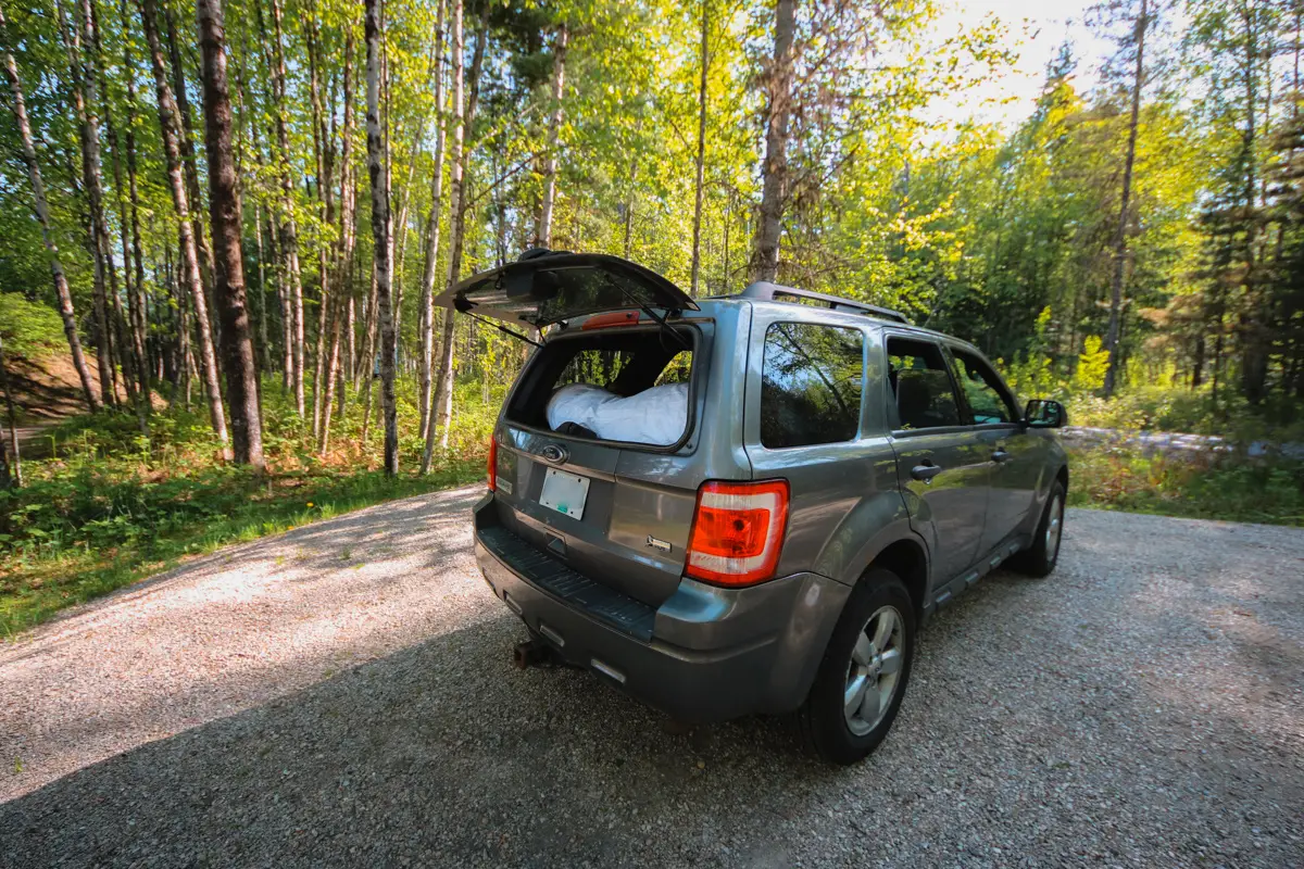 Back hatch of a Ford Escape is open at a campsite at Blanket Creek Provincial Park. Green trees and large gravel pad.