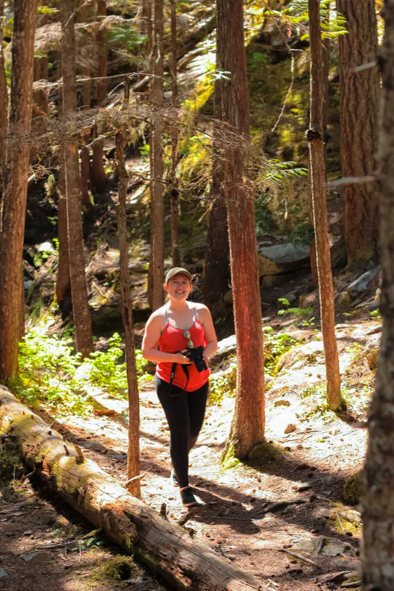 Woman in red tank top smiles at the camera while holding a camera on a dirt hiking trail in a forest.