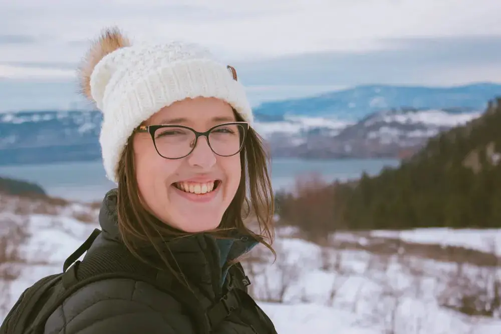 Women with a toque smiles at the camera in the winter. Kal Lake is visible in the background.