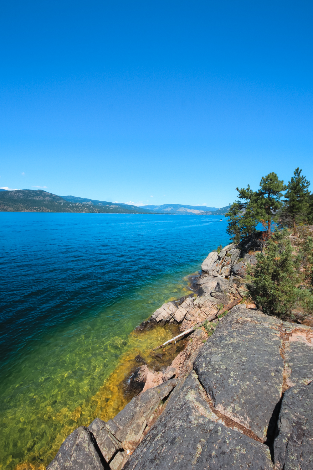 Rocky shorelines on Okanagan Lake. Water is a beautiful blue and green with a blue sky. No clouds.