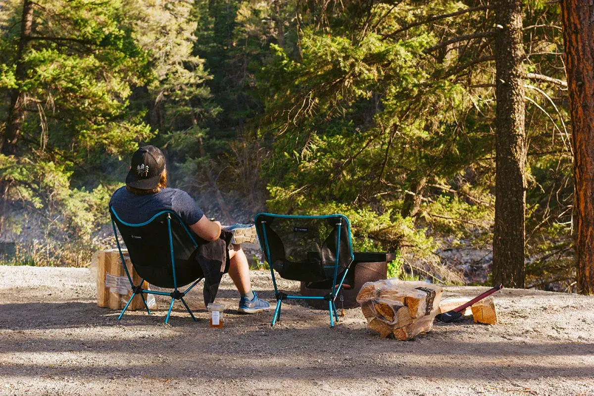 Man sits in a camping chair next to a campfire on a sunny day