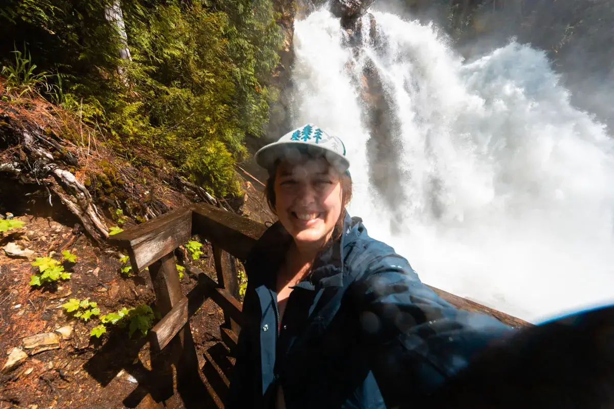 A woman wearing a hat and rain jacket takes a selfie in front of Rainbow Falls near Cherryville, BC.
