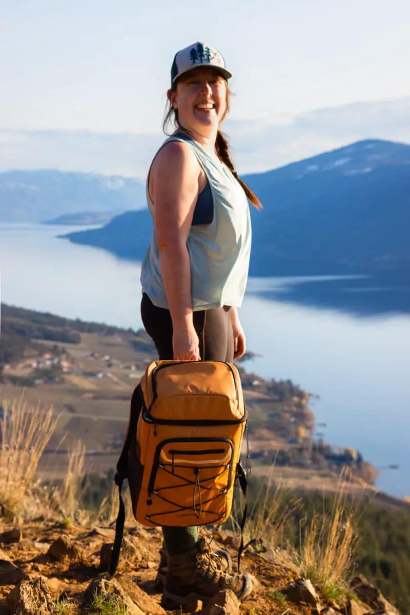 Woman holds the EVERFUN 30-can cooler backpack with lake views in the background.