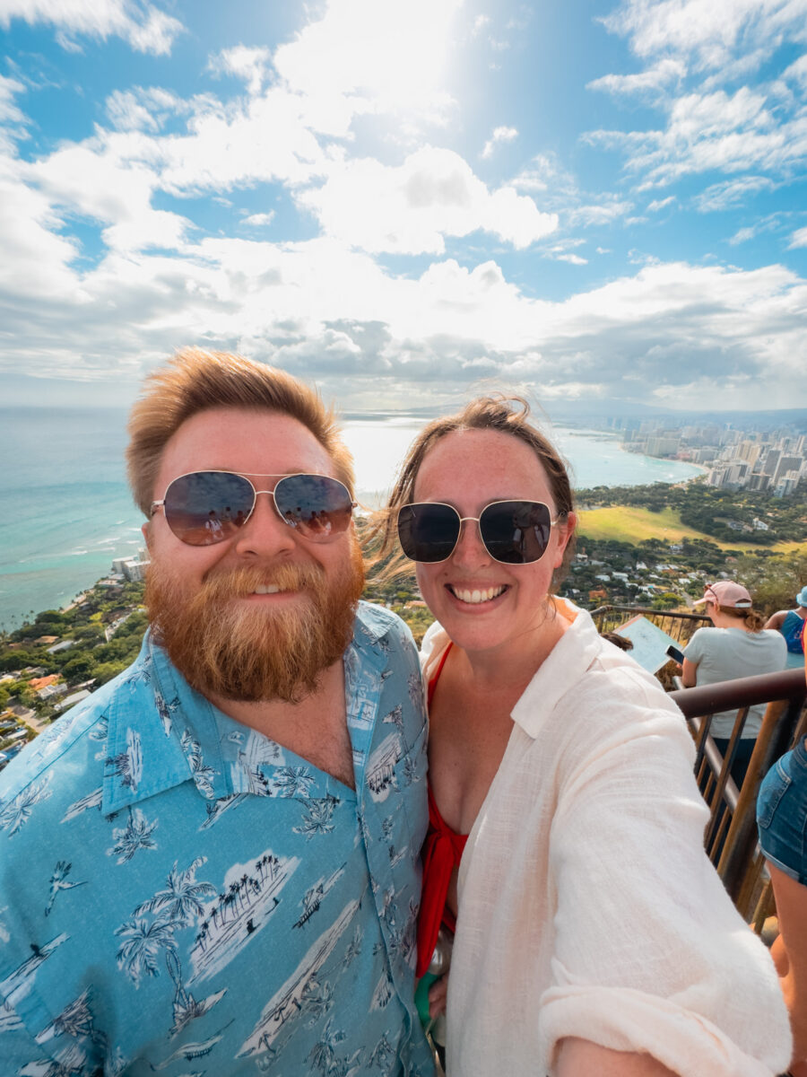 Selfie of a man and woman at the summit of Diamond Head with Waikiki in the distance.