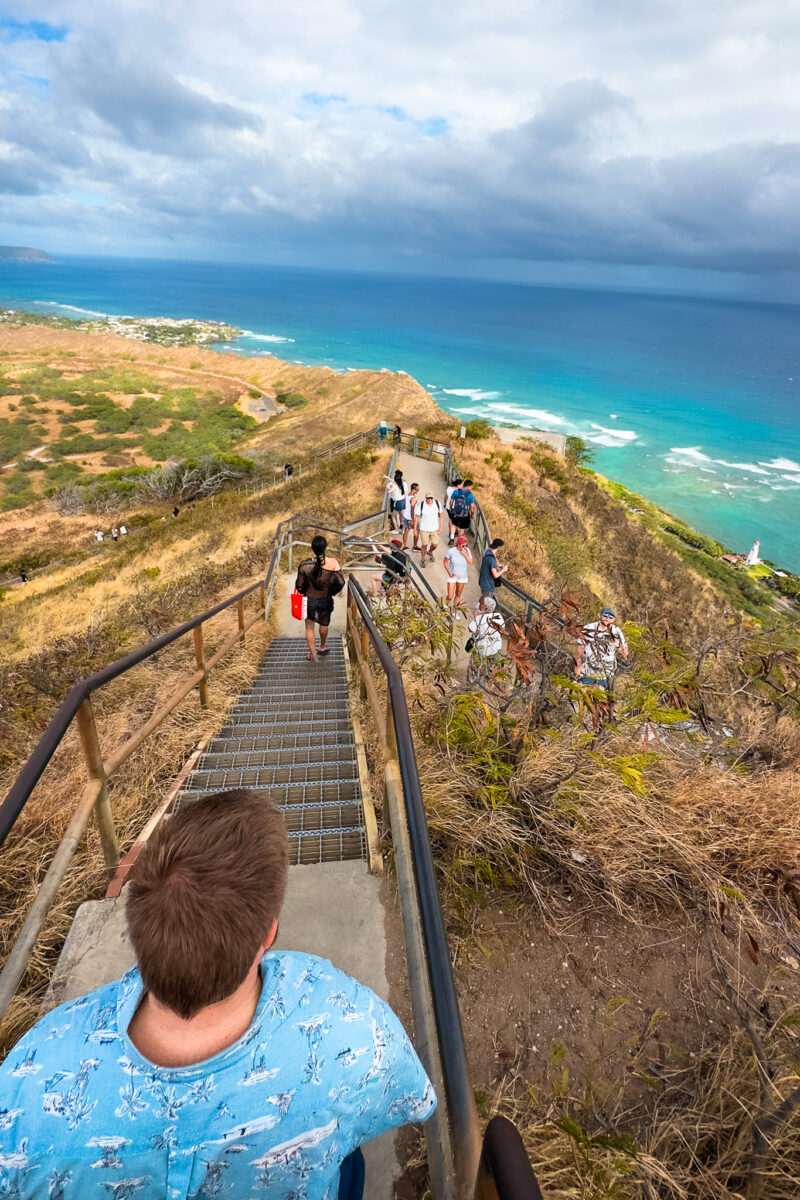 Man walks down a set of stairs with lots of people below. View of the Pacific Ocean and Diamond Head crater wall in the distance.