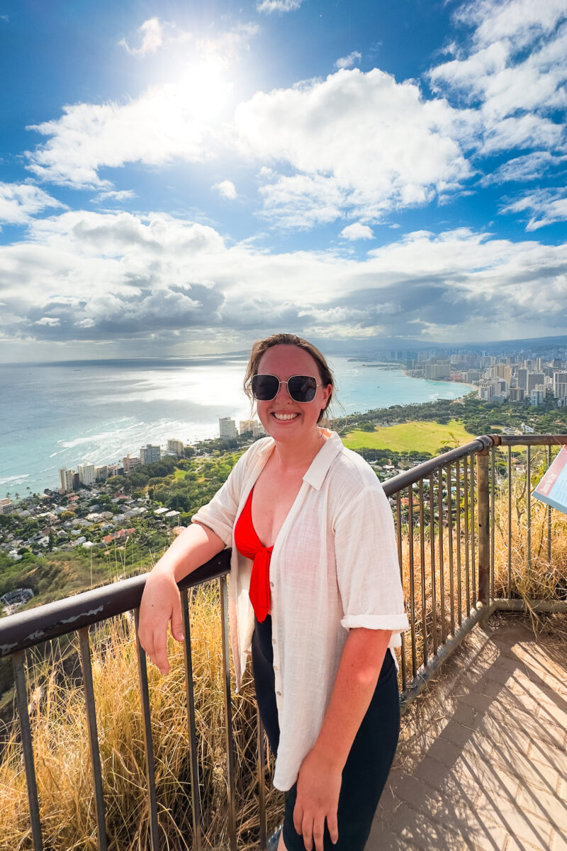 Woman leads against a guardrail at the summit of Diamond Head. Waikiki and the Pacific Ocean are in the background.