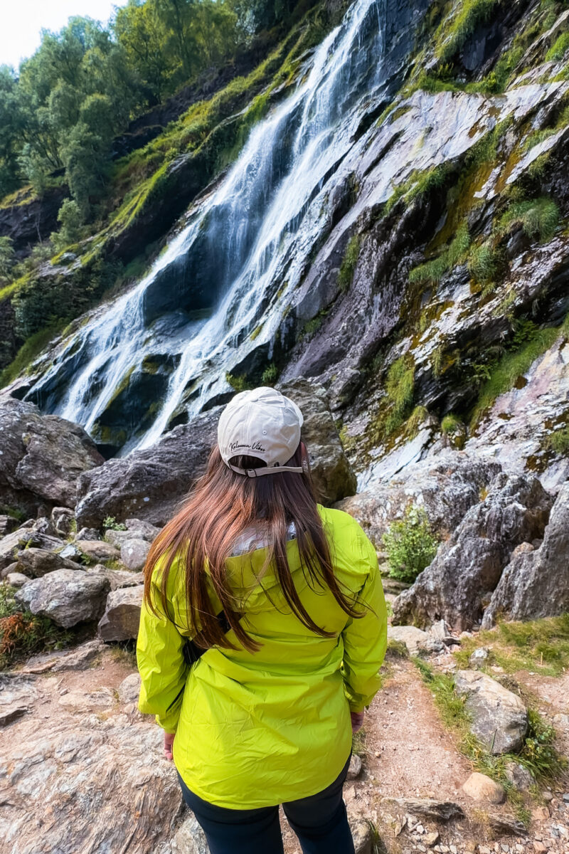 A woman stands at the bottom of a large waterfall.