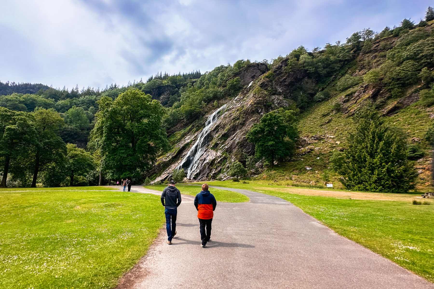 Two men walking on a paved trail towards Powerscourt Waterfall.