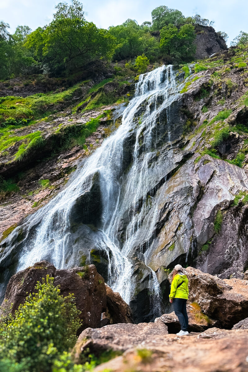 Woman in green waterproof jacket stands at the bottom of Powerscourt Waterfall.