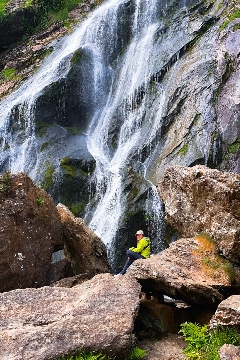 Woman sits at the base of a large waterfall.