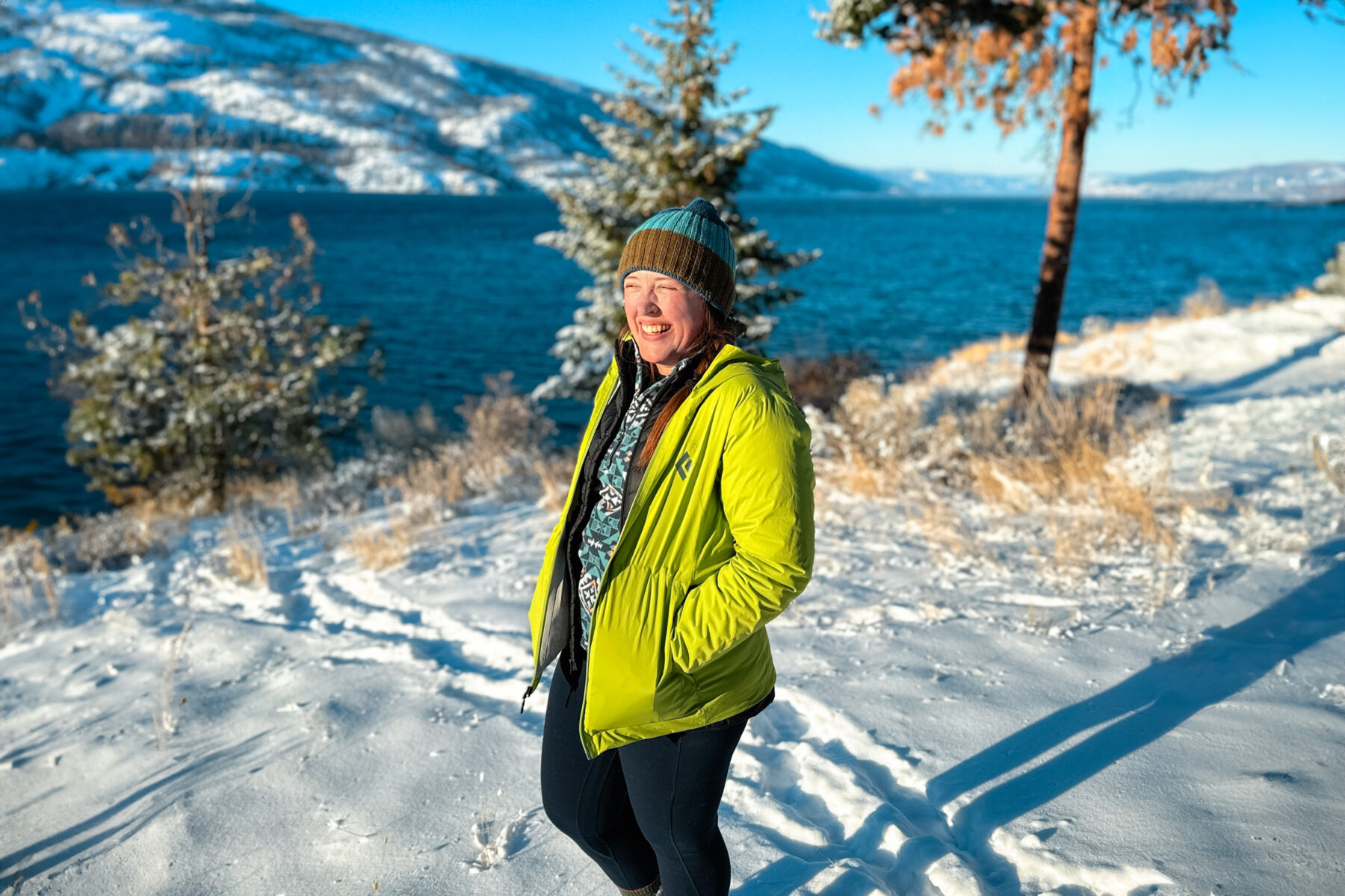 Woman in a green jacket standing on a snowy field in front of Okanagan lake on a sunny day.