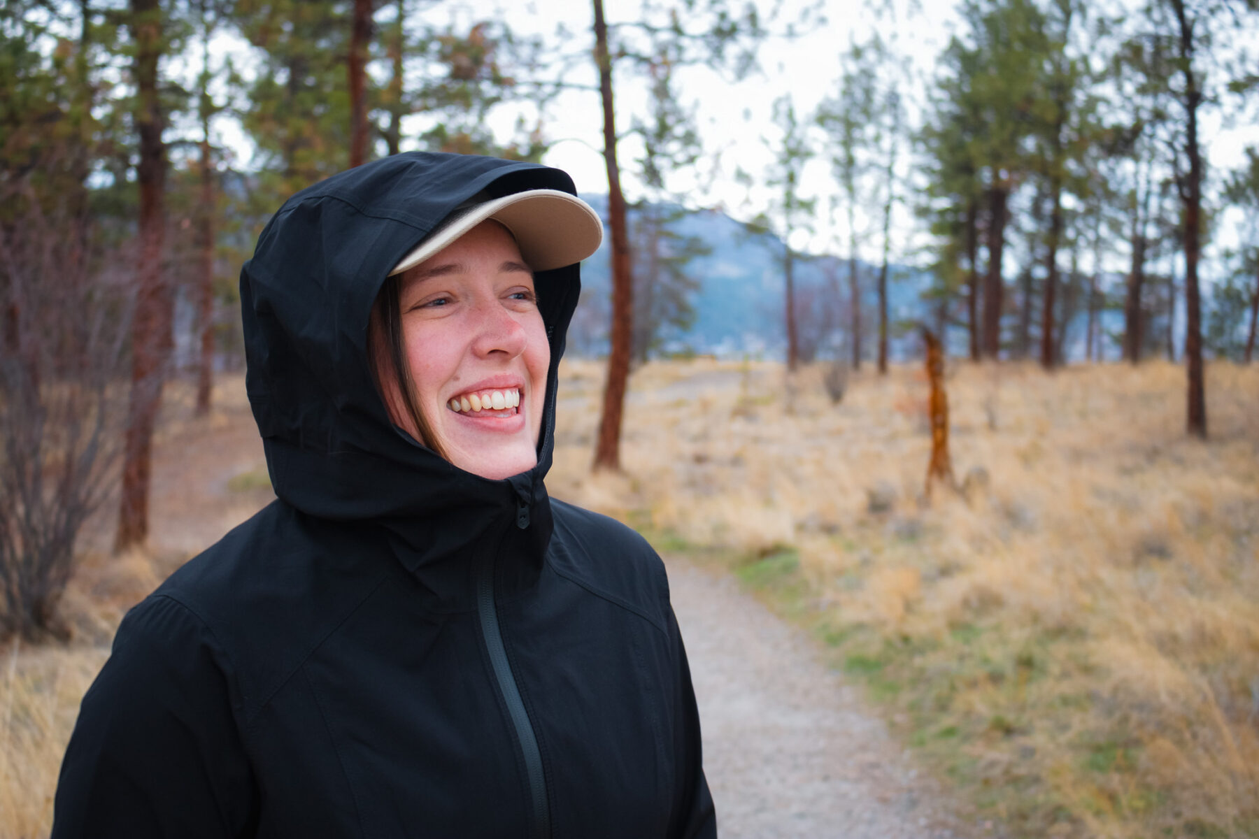 Woman smiling and laughing with a hat underneath the rain jacket's hood.