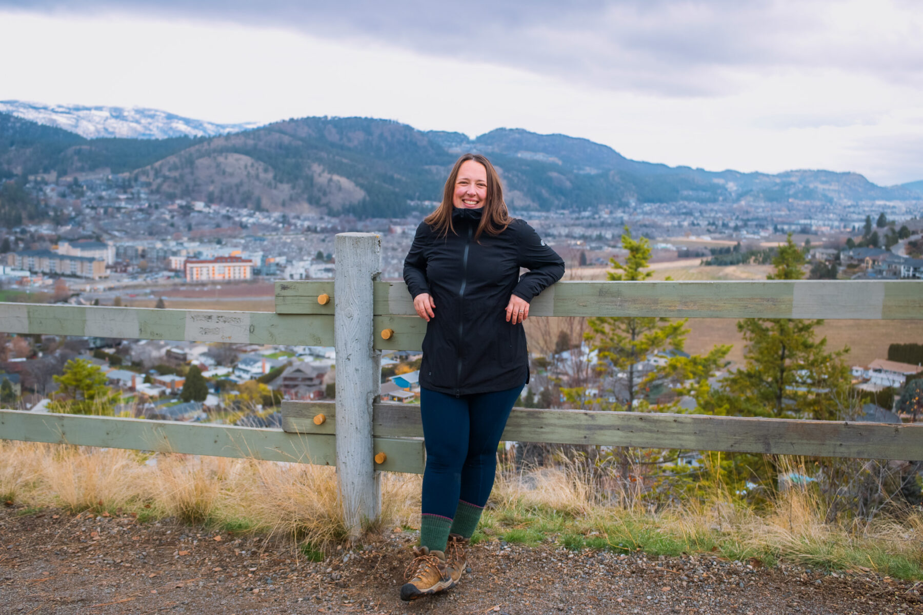 Woman leans against a wooden fence on a rainy day, wearing a black rain jacket.