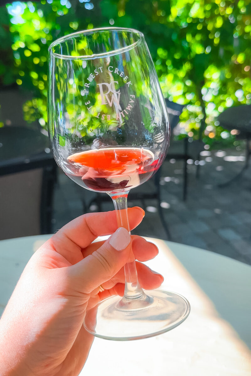 Woman holding a wine glass in front of a vine covered terrace at Priest Creek Winery in Kelowna.