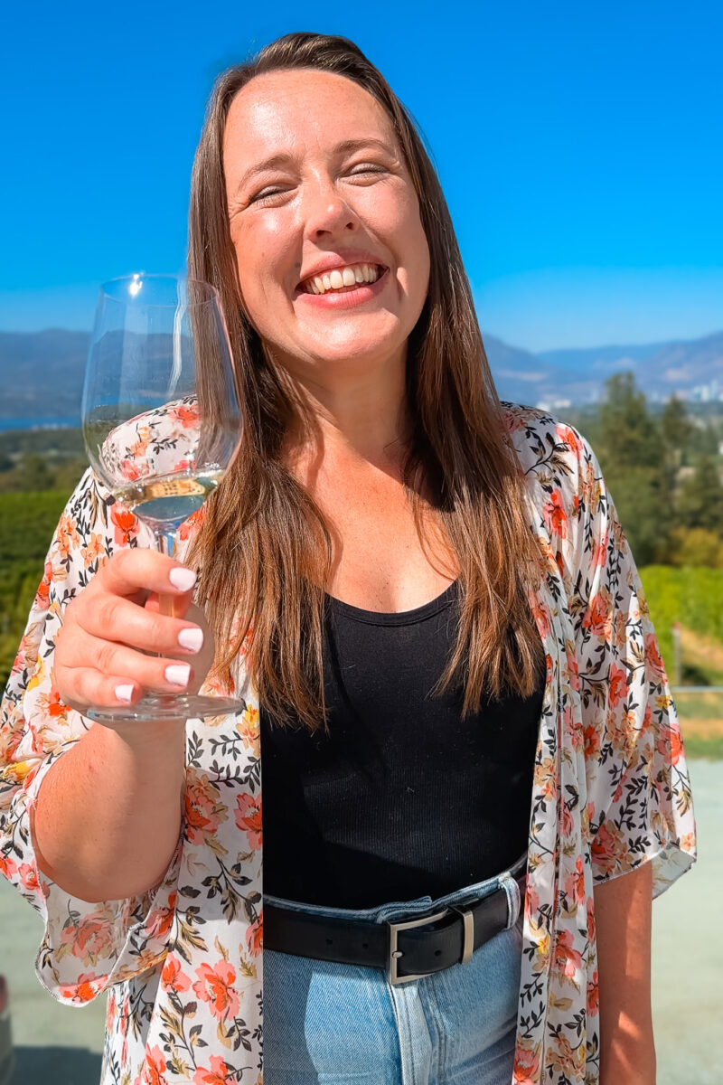 A young woman holding a wine glass and smiling on a sunny day in Kelowna.