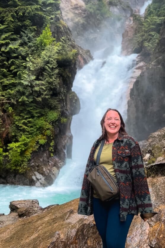 Smiling woman wearing the ALAPAKA Flight Sling bag while standing in front of a waterfall in Canada.